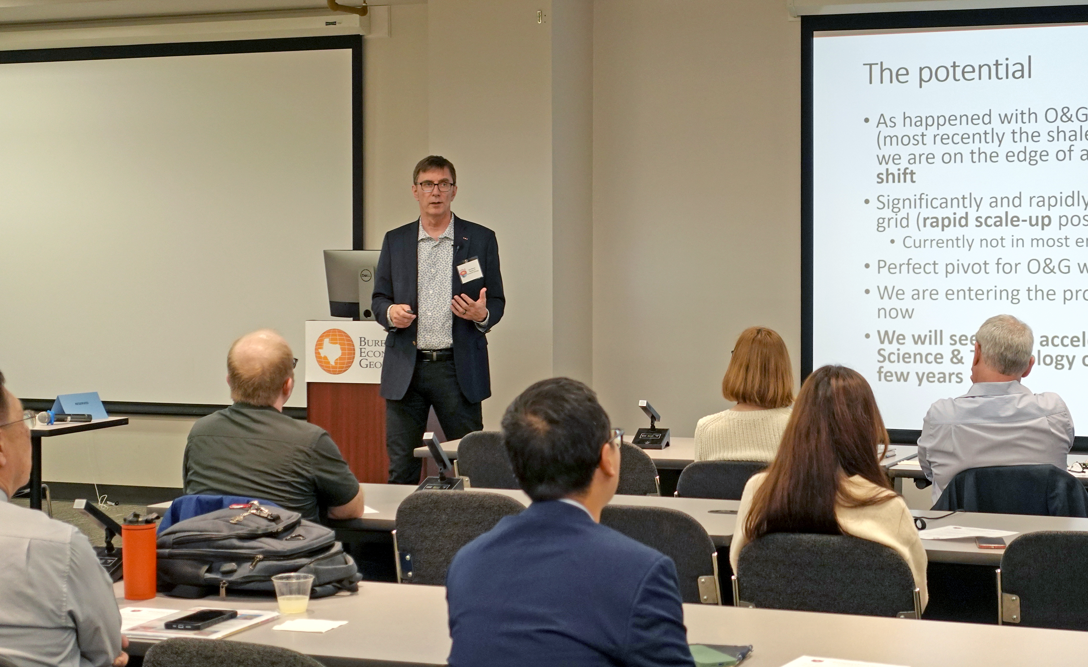 A researcher (Dr. Ken Wisian) stands in front of an attentive audience, presenting a slideshow about research potential for the consortium.