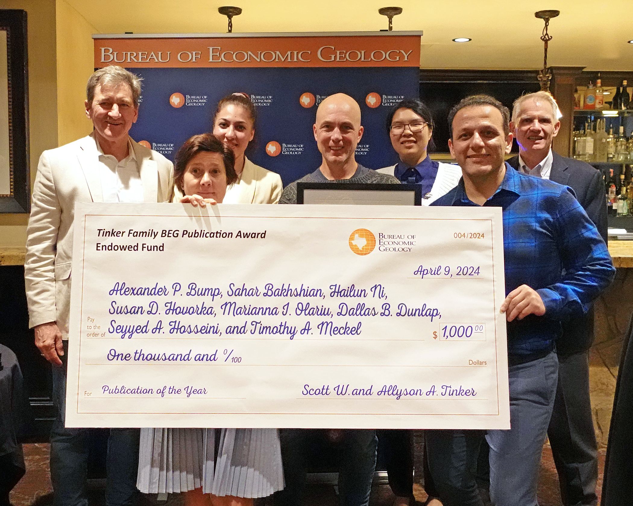 Two presenters and five research award recipients pose for a photo with a large check for $1000 in front of a "Bureau of Economic Geology" patterned step-and-repeat.