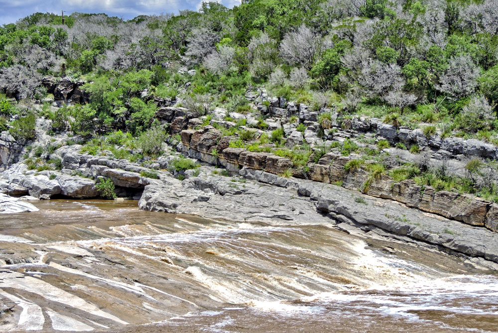 The powerful erosional forces of the river have exposed the dipping Marble Falls beds. Flat-lying Cretaceous unconformity lie in the trees above.
